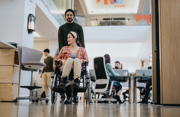 Caring colleague pushing wheelchair of female coworker in modern office.