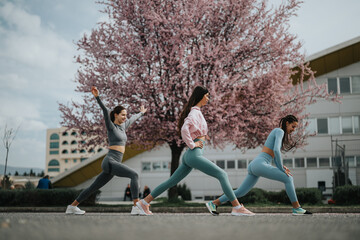 Three women engage in fitness stretching exercises outdoors with blooming cherry blossoms in the...