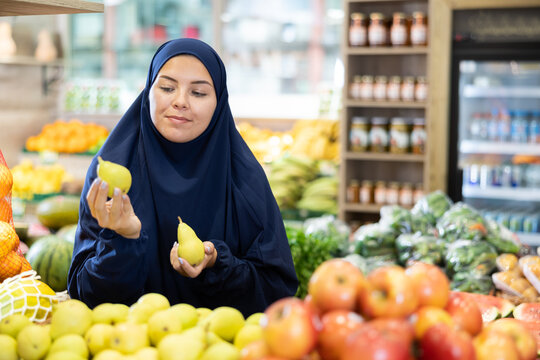 Happy smiling girl in paranja making purchases in supermarket, choosing fresh ripe pears