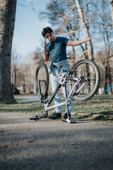 A young adult appears perplexed and calling for assistance with his bicycle on a sunny day in the park.