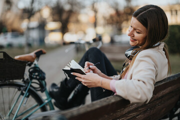 A cheerful young female enjoys journaling on a bench in the park beside her vintage bicycle,...