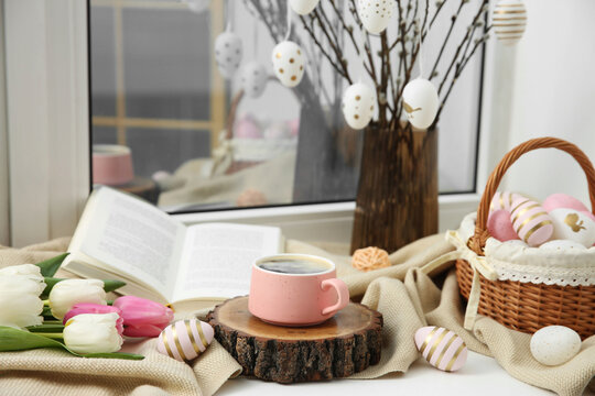 Easter decorations. Cup of coffee, painted eggs, tulips and book on windowsill