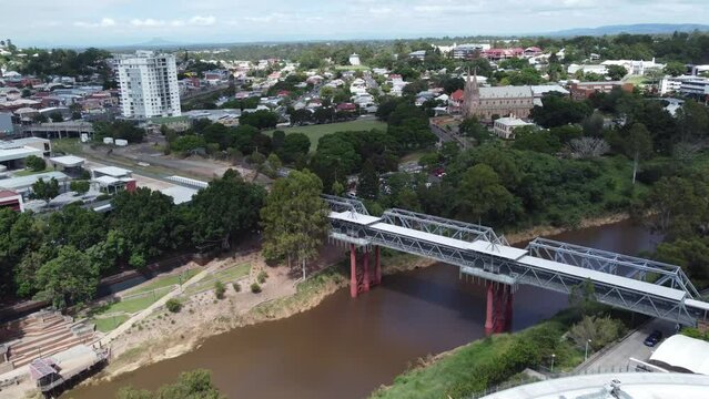 Aerial view of a pedestrian bridge over a brown river approaching a green field and a large Catholic Church in Australia