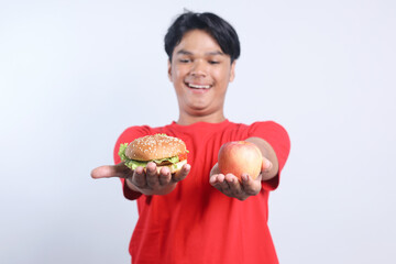 Cheerful young Asian man holding hamburger and apple fruit choosing between healthy or unhealthy food against white background.