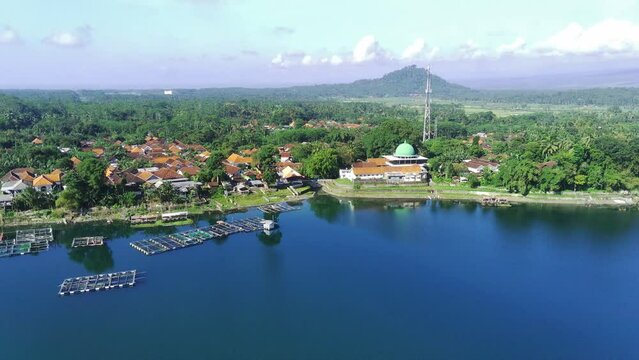 An aerial view of a serene lake with a white mosque with a dome and tall minaret in the background. Lush greenery surrounds the lake. Near Ranu Pakis, Lumajang, East Java, Indonesia.