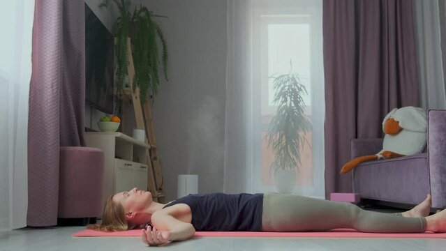 Attractive blonde woman working out at home, lying in Shavasana, resting after yoga practice, meditating, breathing.