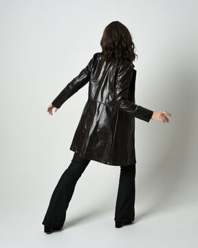 Full length portrait of brunette woman wearing long leather trench coat and black boots. Standing pose walking away from camera, facing backwards. Isolated silhouette on white studio background.