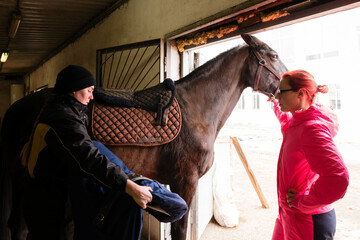Two people saddling a brown horse in stable.