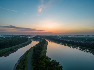 Captured at twilight, this serene aerial image presents a meandering river flowing gently past a small riverside community. The placid water reflects the soft colors of the sunset sky, ranging from