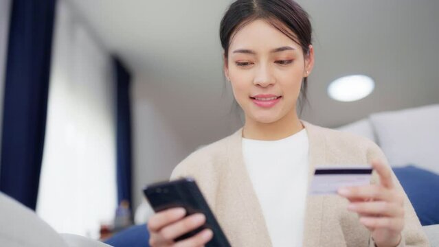 Young asian woman sitting on sofa in living room, makes online banking payments through the internet from bank card on smartphone. Shopping online on mobile phone with credit card