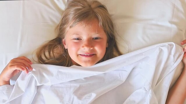 A charming happy little girl with long hair is lies on a white bed under a white blanket and laughs 
