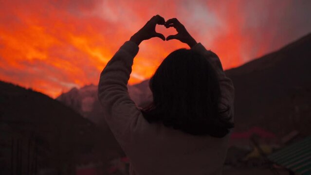 Silhouette shot of girl making heart shape with hands while standing against mountains and sunset sky in Keylong, Lahaul, India. Finger-shaped heart shape. Hands of girl shape of heart. Summer dream. 