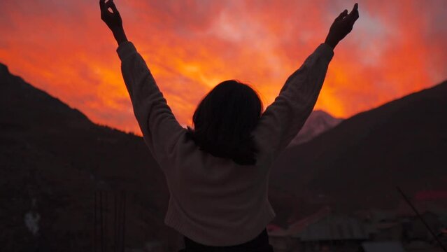 Silhouette shot of Girl raise her arms against sunset sky and snowy mountain peaks in Keylong, Lahaul, India. Young woman standing and relaxing on mountain. Happy girl silhouette at sunset