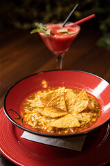 mexican tortilla soup served in red dish in restaurant with red cocktail on dark background