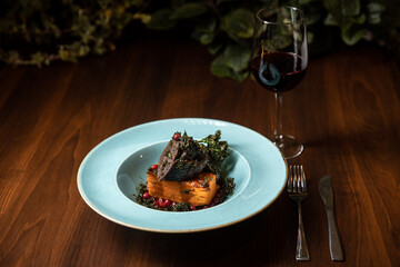 cooked meat with potato lasagna served in blue dish in restaurant with glass of wine on dark background