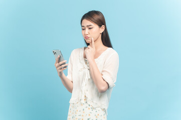 Beautiful young asian woman in white dress with flower pattern using smartphone with unsure feeling isolated on blue background