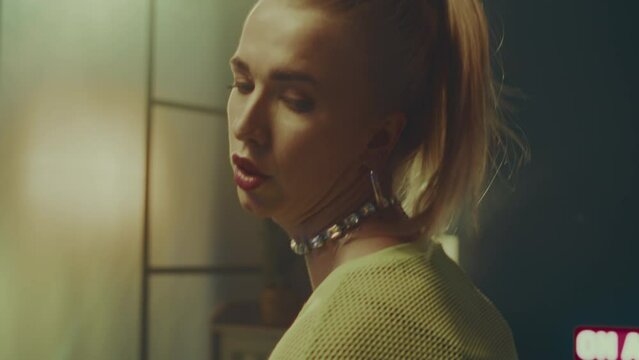 Tilting up shot of seductive young Caucasian blonde woman wearing fishnet tights with high waist mini shorts and neon yellow semi sheer long sleeve crop top performing vogue dance movements indoors
