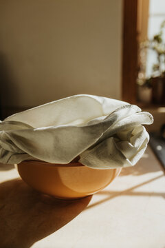 Bread proofs in bowl with tea towel on kitchen counter in sunshine