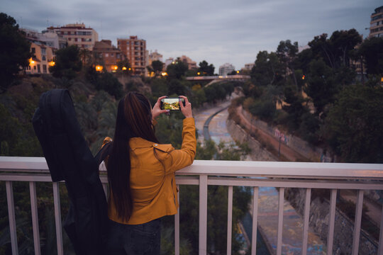 Woman with a guitar taking picture of river from urban bridge at dusk