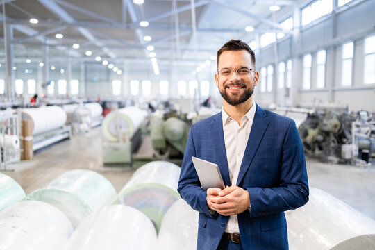 Caucasian factory manager or CEO holding tablet computer and checking production results in industrial plant while employees working in background.