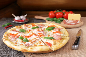 On a wooden table against a log wall is a traditional pizza with ham and tomatoes next to a cutting board with ingredients. 