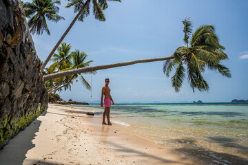 Mężczyzna w kąpielówkach na rajskiej plaży pod palmą - obrazy, fototapety, plakaty