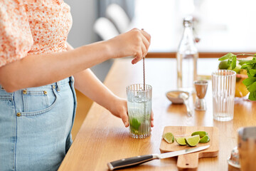 culinary, drinks and people concept - close up of woman with glass and spoon making lime mojito cocktail at home kitchen