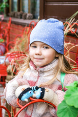 A young girl with blonde hair smiles in a blue hat with white polka dots, a colorful jacket, and...