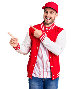 Young handsome man wearing baseball jacket and cap smiling and looking at the camera pointing with two hands and fingers to the side.