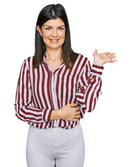 Beautiful brunette woman wearing striped shirt smiling cheerful presenting and pointing with palm of hand looking at the camera.