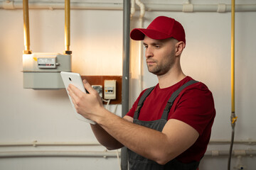 A skilled technician, wearing a red cap and overalls, is inspecting a heating system in the...
