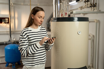 A young woman examines a non-operational boiler with concern, holding a phone in her hand, likely...