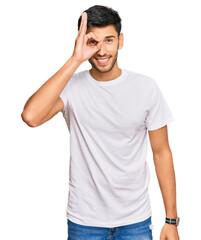 Fototapeta premium Young handsome man wearing casual white tshirt doing ok gesture with hand smiling, eye looking through fingers with happy face.