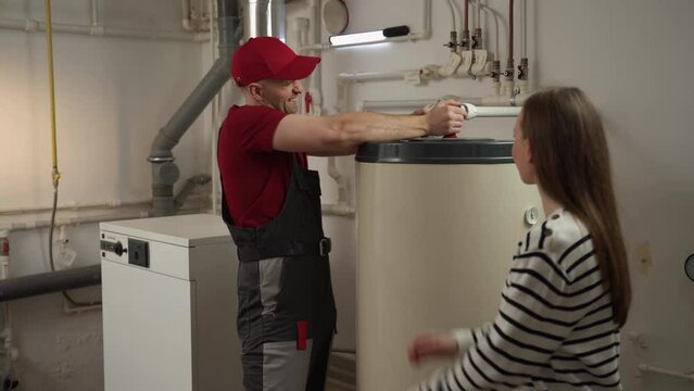 A knowledgeable plumber in a red cap and work uniform is engaging with a homeowner, discussing the maintenance of a water heater in a residential basement. He is providing explanations and