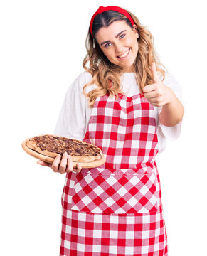 Young caucasian woman wearing apron holding pizza smiling happy and positive, thumb up doing excellent and approval sign