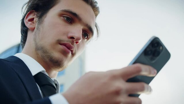 Confident businessman near a business center with a cell phone in hands. Successful  young businessman in a blue suit is looking at a smartphone. Businessman is typing message on a mobile phone.