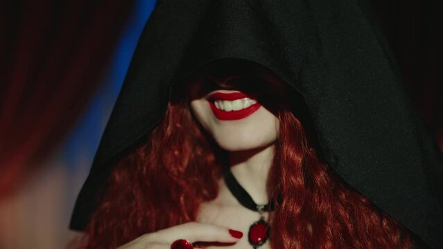 Close-up portrait mouth red lips beauty face sexy vampire woman happy smiling shows white long false fangs. art halloween party make-up redhead. Gothic lady fantasy girl hiding face black hood dress