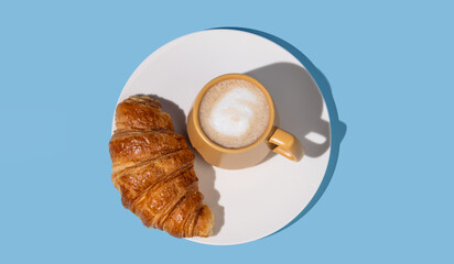 Flat lay of coffee cup and fresh croissant on a white plate on a blue background.