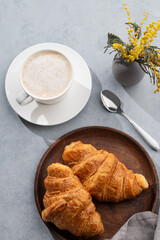 A cup of aromatic cappuccino coffee and fresh croissants in a wooden plate on a blue background with yellow flowers.