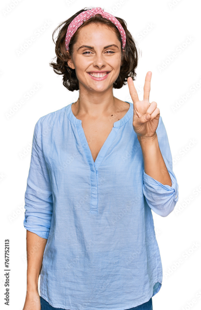 Wall mural Young hispanic woman wearing casual clothes smiling with happy face winking at the camera doing victory sign. number two.