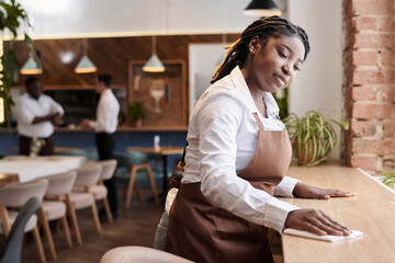 Medium shot of young African American waitress wiping table at work in modern restaurant, copy space - obrazy, fototapety, plakaty