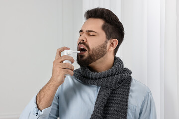 Young man with scarf using throat spray indoors