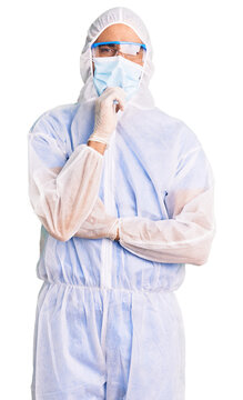 Young hispanic man wearing doctor protection coronavirus uniform and medical mask with hand on chin thinking about question, pensive expression. smiling with thoughtful face. doubt concept.