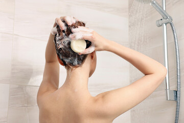 Young woman washing her hair with solid shampoo bar in shower, back view