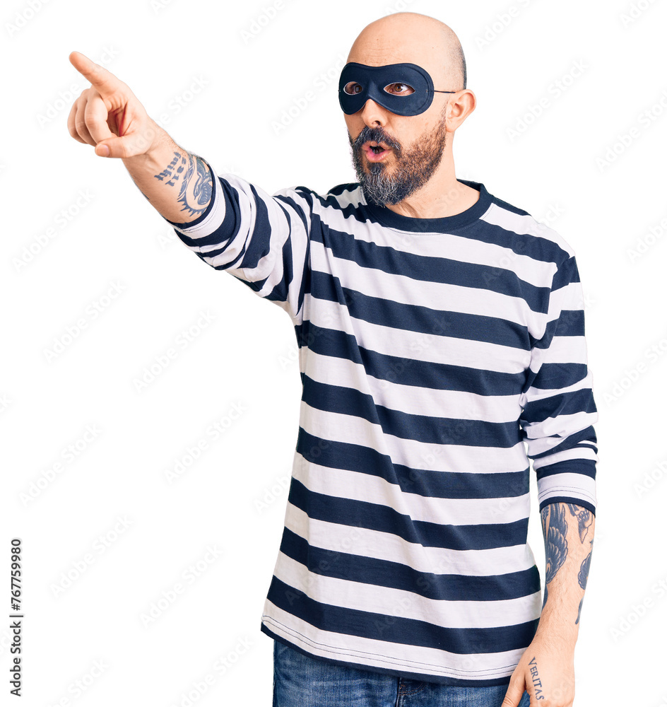 Sticker Young handsome man wearing burglar mask pointing with finger surprised ahead, open mouth amazed expression, something on the front