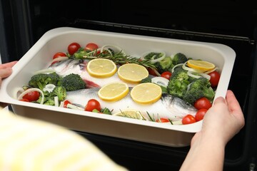 Woman putting baking dish with raw fish and vegetables into oven, closeup