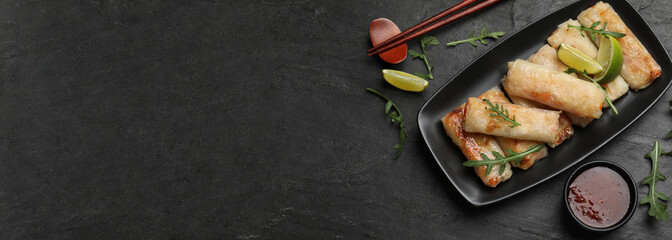Delicious fried spring rolls, sauce and chopsticks on black table, flat lay. Banner design with space for text