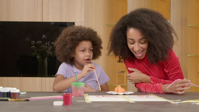 Funny cute school age African American girl trying to blow paints on paper with all her might, expressing confusion and positive emotions while straw painting together with cheerful mother.