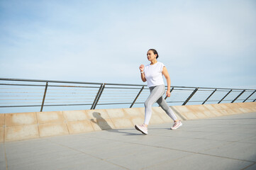 Full length portrait of a young adult woman in sports clothing, running on the promenade long the bridge