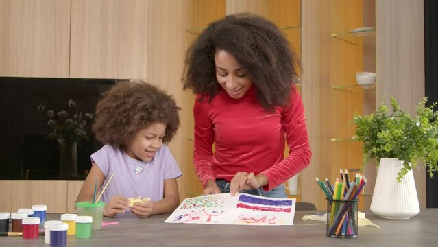 Joyful cute school age African American girl and cheerful attractive black mother looking and enjoying self painted picture on paper, giving high five gesture , expressing happiness and positivity.